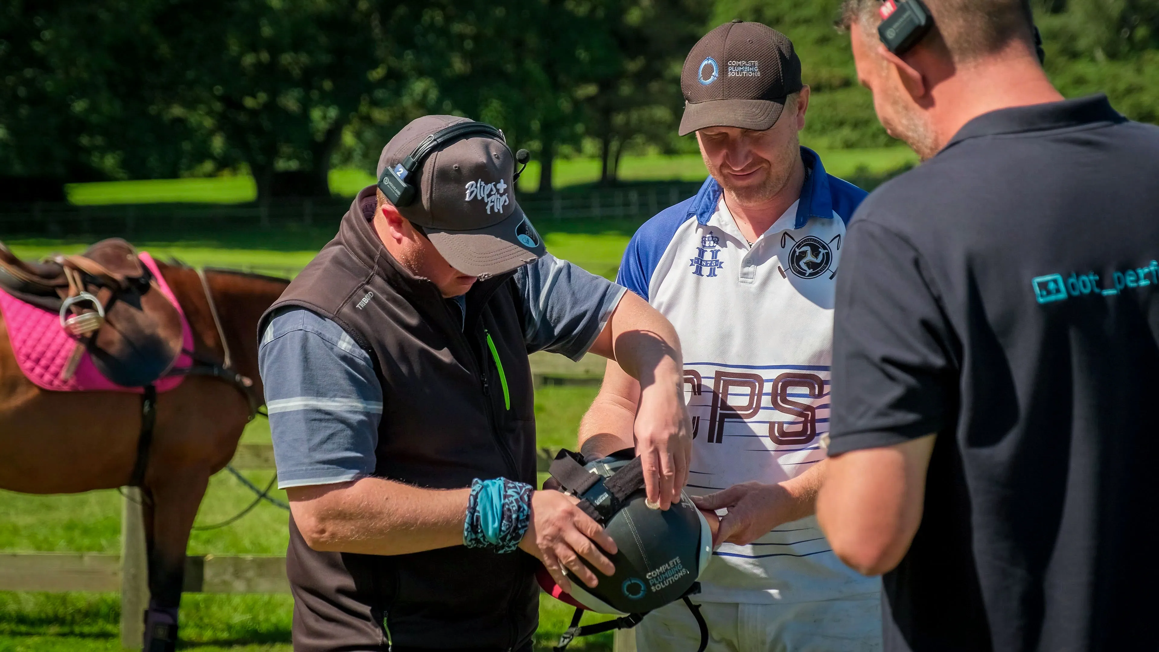 Three men are outdoors examining a helmet. One adjusts it while the others watch. A brown horse with a pink saddle stands nearby. The mood is focused.